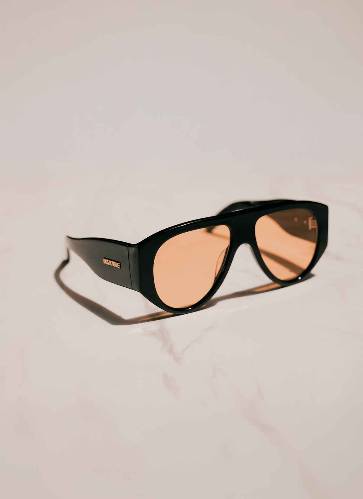 Black oversized sunglasses with warm amber-tinted lenses, resting on a light beige marble surface with a soft shadow.