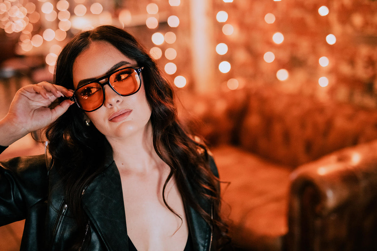 A woman with long dark hair wearing black-framed sunglasses with orange-tinted lenses and a black leather jacket, posing in a warmly lit indoor setting with bokeh string lights in the background.