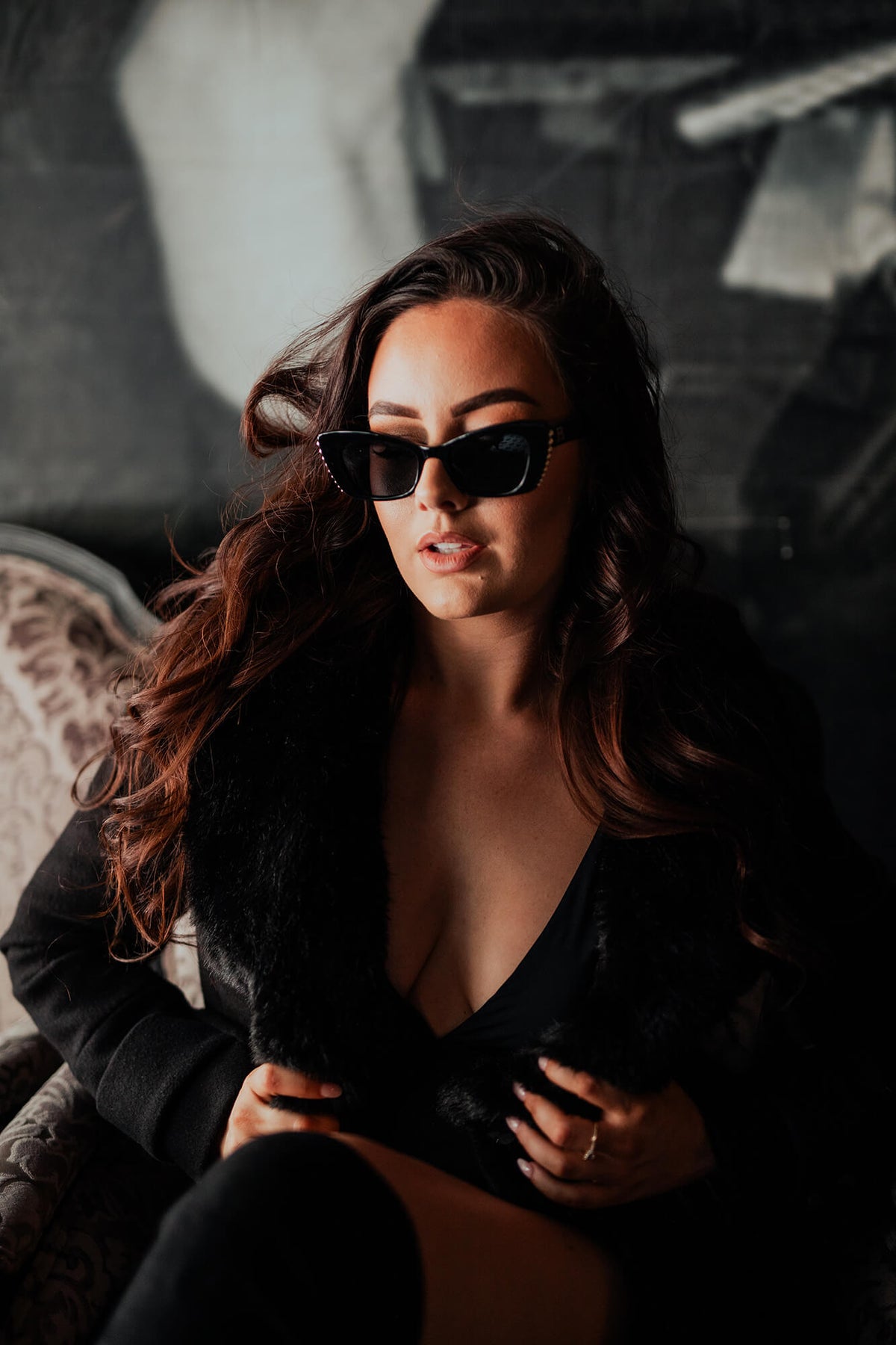 A woman with long wavy brown hair wearing black cat-eye sunglasses, a black faux-fur collar jacket, and black thigh-high stockings, seated in a dimly lit interior setting.