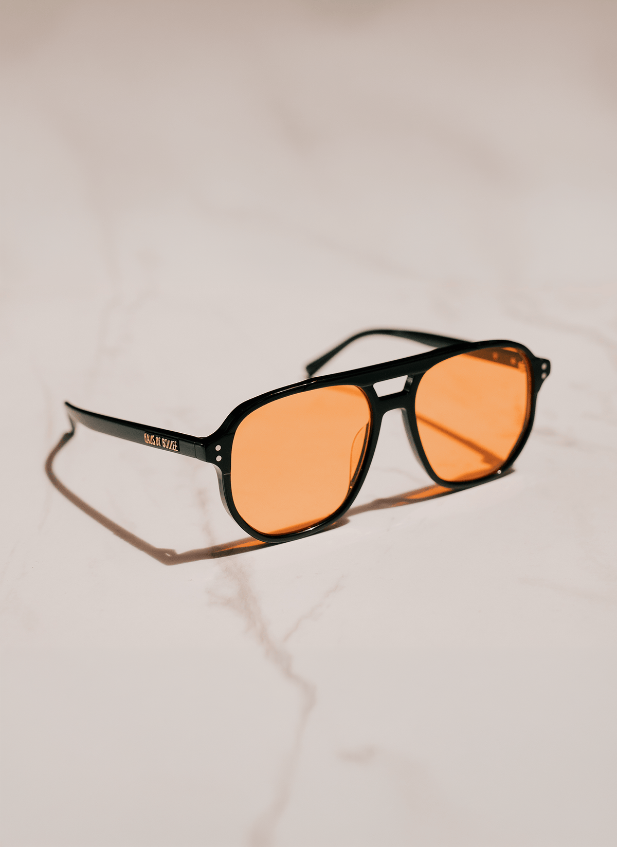 Black square aviator-style sunglasses with orange tinted lenses, resting on a light beige marble surface.