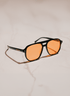Black square aviator-style sunglasses with orange tinted lenses, resting on a light beige marble surface.