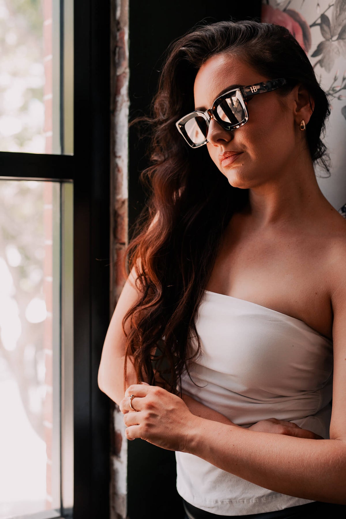 A woman with long wavy brown hair wearing black-framed square sunglasses and a white strapless top, standing indoors near a window.