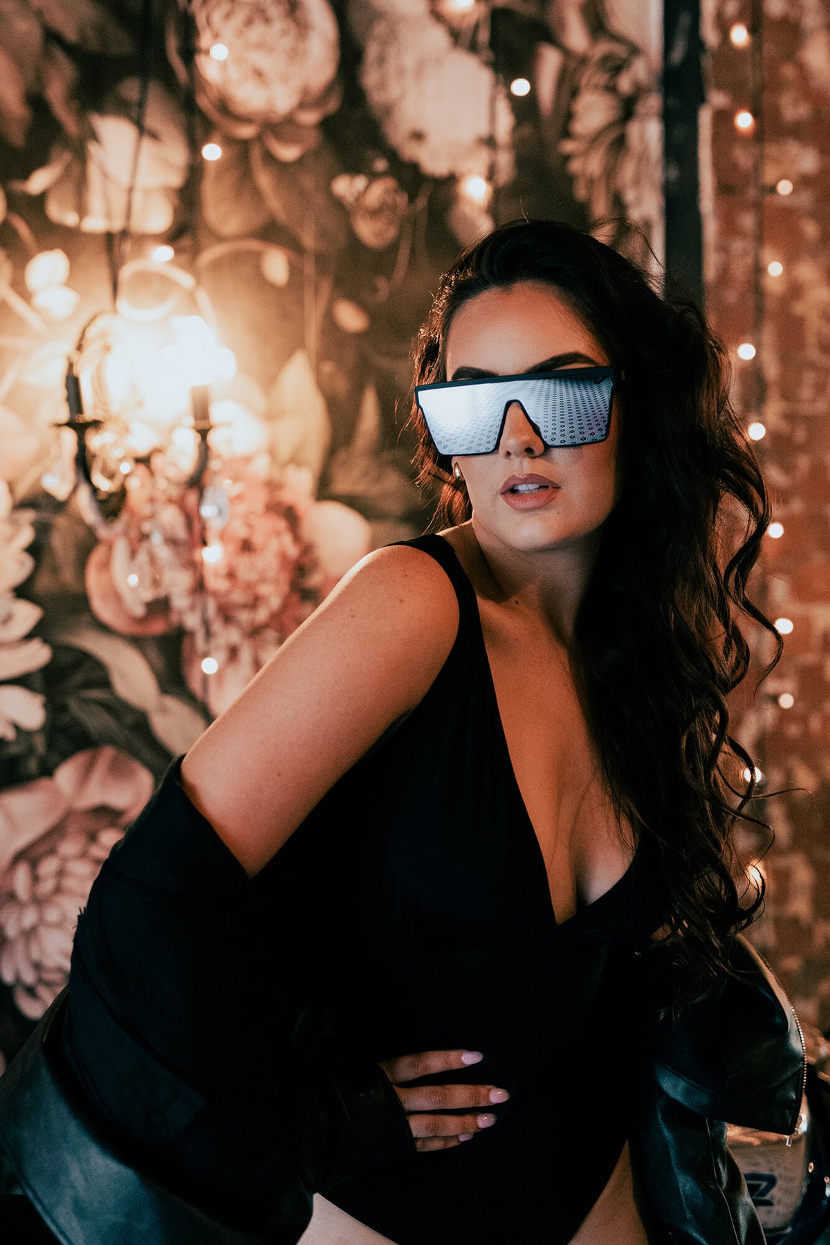 A woman with long dark wavy hair wearing futuristic silver visor sunglasses and a black bodysuit, posing in front of a warm-toned floral wall with hanging lights.