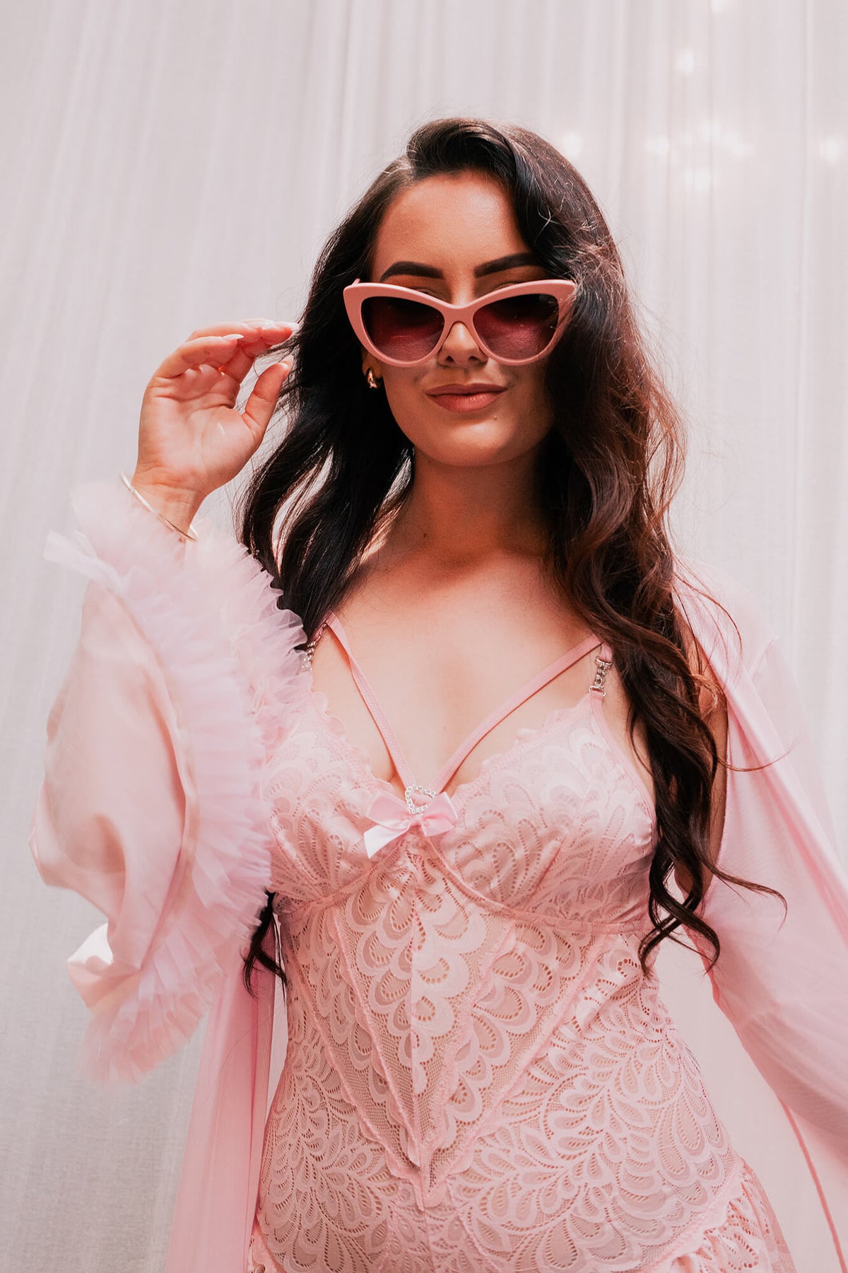 A woman with long wavy dark hair models a light pink lace bodysuit with a matching sheer ruffled robe and pink cat-eye sunglasses against a soft white curtain background.