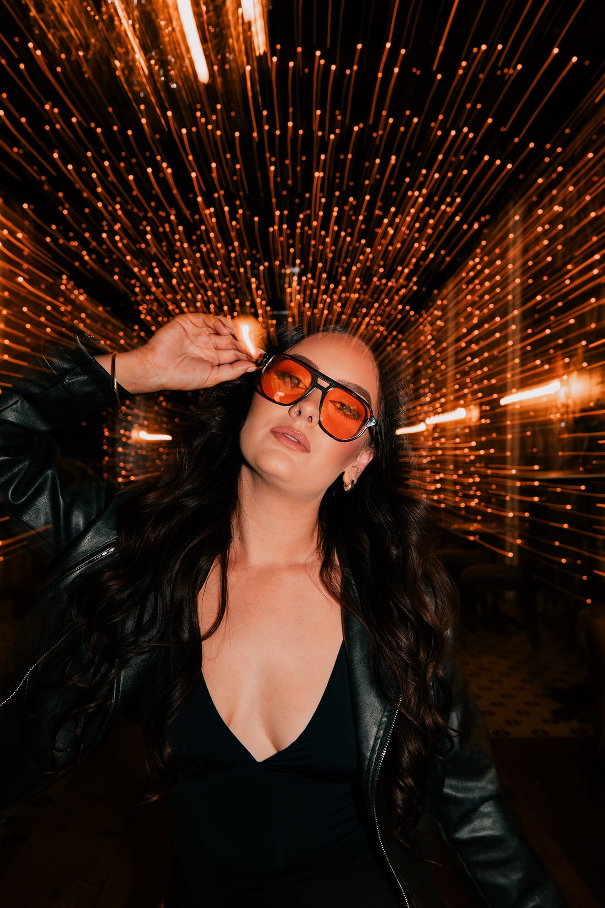 A woman wearing black sunglasses with orange lenses and a black leather jacket poses in front of a tunnel of glowing orange light streaks.