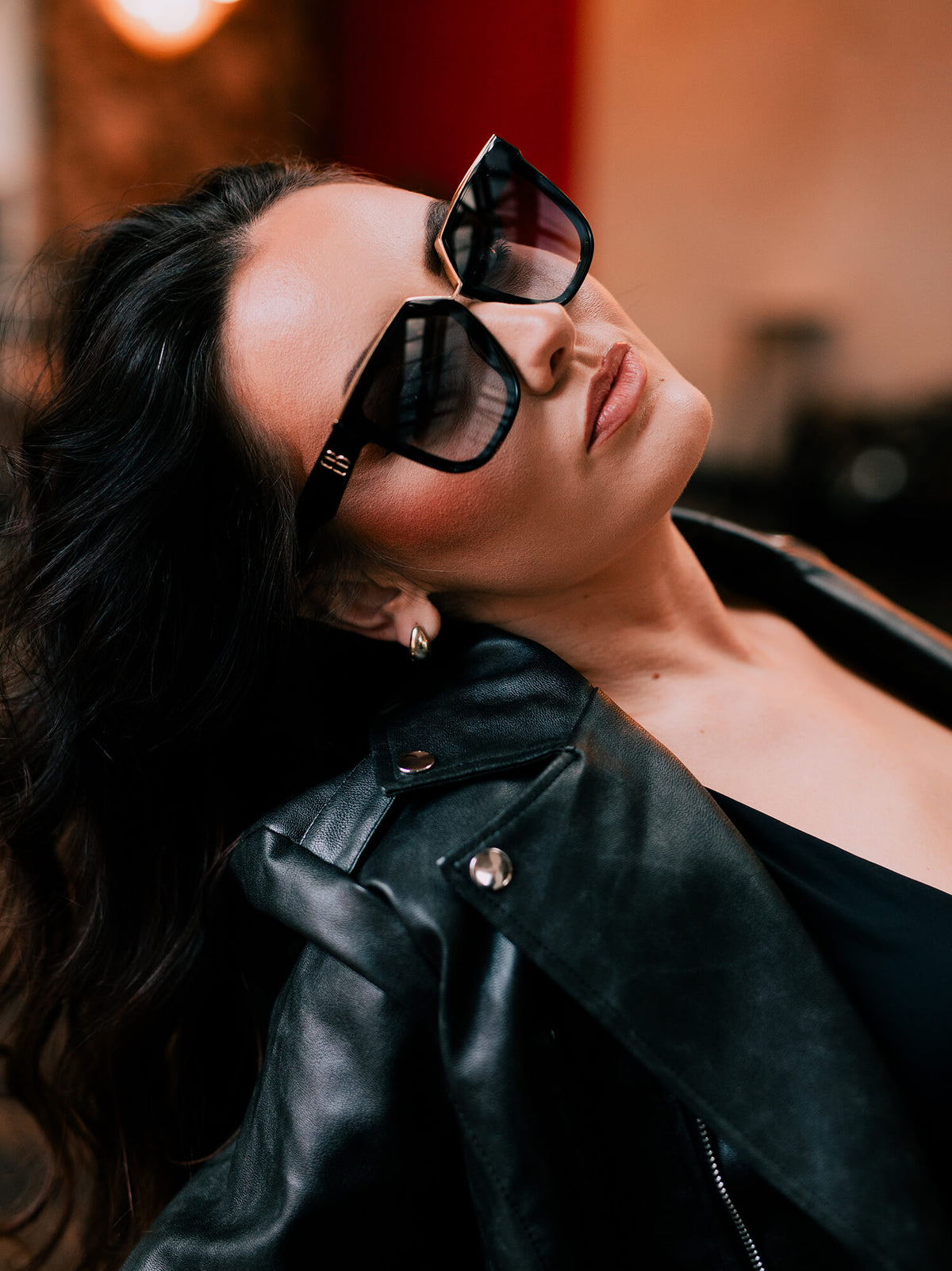 A close-up lifestyle shot of a woman wearing oversized black sunglasses with gold-toned metal frames and a black leather jacket, set against a warm, blurred indoor background.
