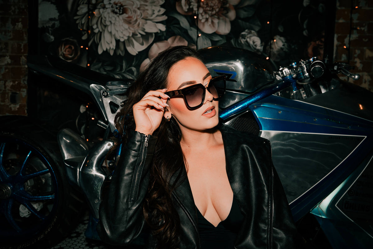 A woman wearing black sunglasses and a black leather jacket poses in front of a blue and black motorcycle, with a dark floral wall backdrop.