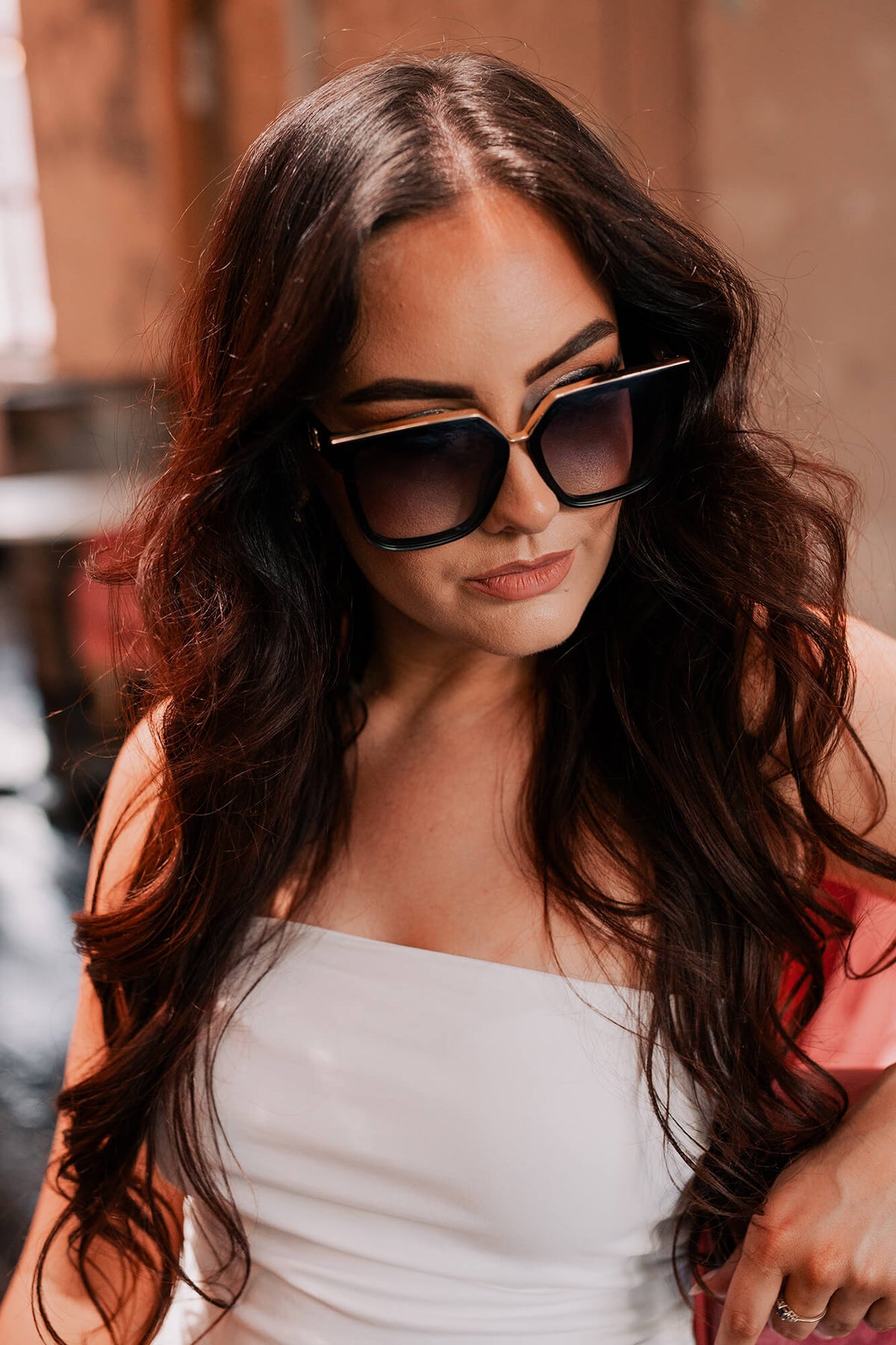 A woman with long, wavy dark brown hair wearing chunky black cat-eye sunglasses with gold metal trim and a white strapless top, set against a warm, blurred background.