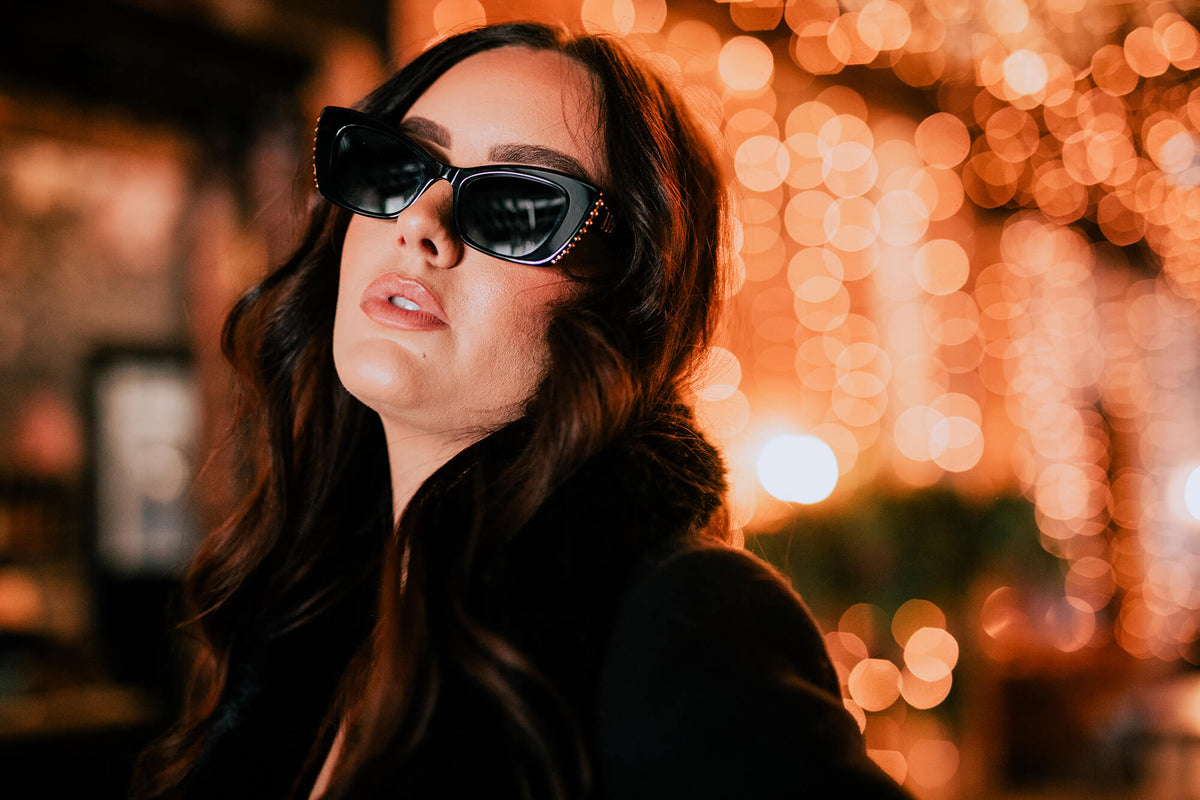 A woman with long wavy brown hair wearing black cat-eye sunglasses with gold chain accents on the temples, set against a warm, out-of-focus bokeh background of orange lights.