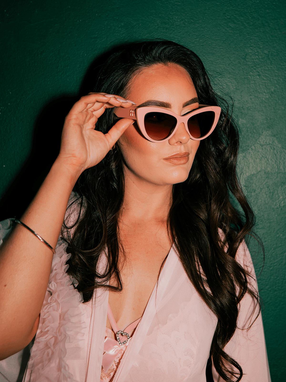 A woman with long dark wavy hair wearing light pink cat-eye sunglasses and a matching light pink robe, posing against a dark green textured wall.