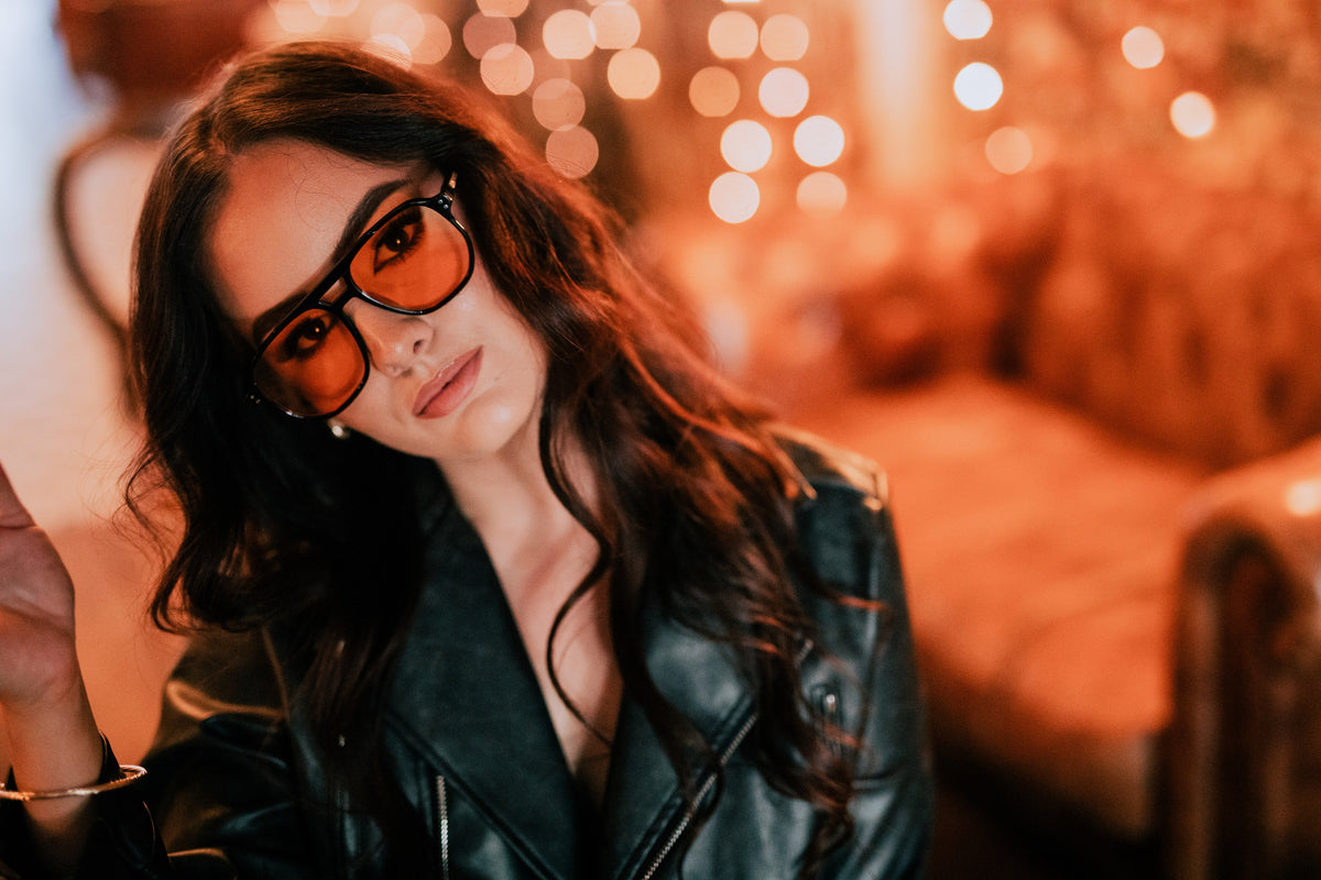Woman wearing a black leather jacket and orange-tinted glasses with a blurred background